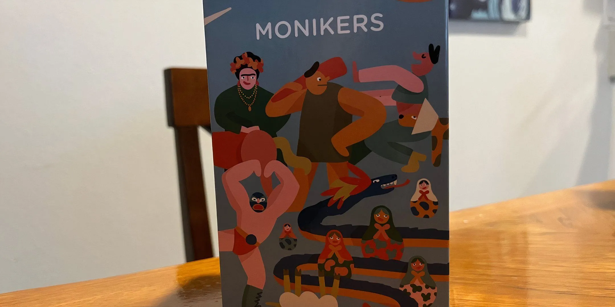 Bìa hộp game Monikers với phong cách minh họa vui nhộn, gợi ý về một trò chơi đoán từ đầy tính sáng tạo và hài hước.